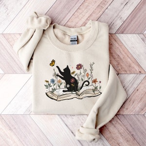 May include: A beige sweatshirt with a black cat silhouette sitting on an open book with flowers and a butterfly. The cat is reaching for the butterfly. The text "Gildan" is visible on the tag.