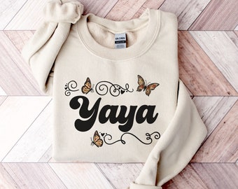 May include: A sand-colored sweatshirt with the word "Yaya" in a black, retro font. The word is surrounded by butterfly graphics and swirls.