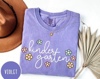 May include: A violet Comfort Colors t-shirt with the word "kindergarten" in white script, decorated with colourful flower graphics. A gold necklace and a brown hat are also in the image.
