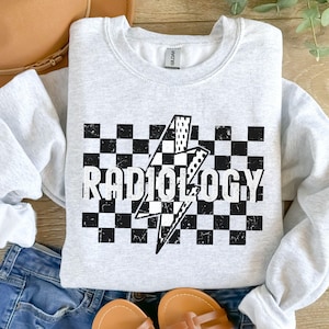 May include: A gray sweatshirt with a black and white checkered pattern and a lightning bolt graphic. The text "RADIOLOGY" is printed in black on the checkered pattern.