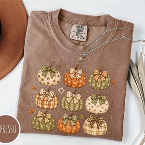 May include: An espresso-colored t-shirt featuring a graphic print of nine pumpkins, each adorned with a patterned bow. The pumpkins display various designs including hearts, stars, and plaid. The shirt has a Comfort Colors label at the neck.