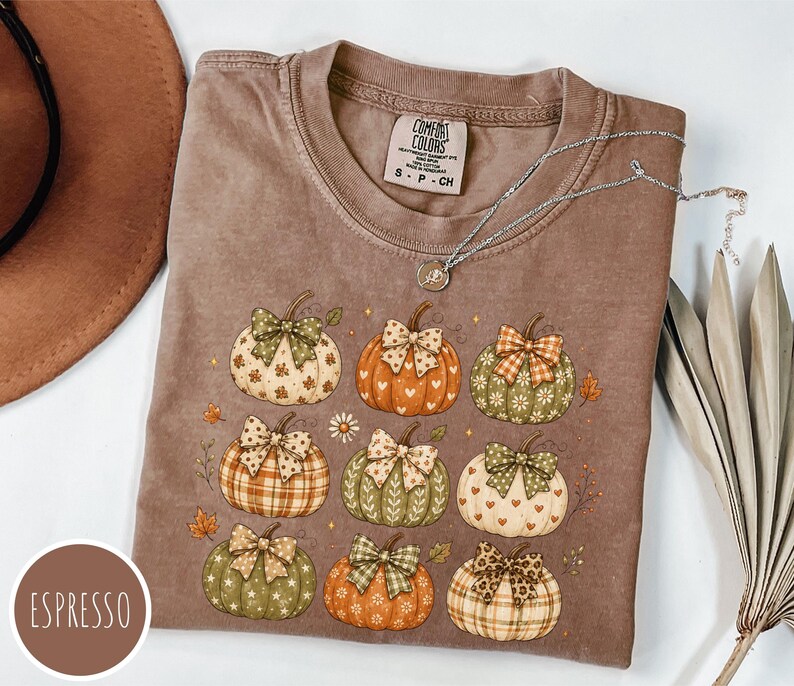 May include: An espresso-colored t-shirt featuring a graphic print of nine pumpkins, each adorned with a patterned bow. The pumpkins display various designs including hearts, stars, and plaid. The shirt has a Comfort Colors label at the neck.