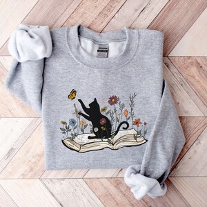 May include: A gray sweatshirt with a black cat sitting on an open book. The cat is reaching for a yellow butterfly. There are colorful flowers around the book and cat.