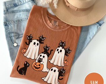 May include: A rust-colored t-shirt featuring a graphic print of white ghosts and black cats interacting with orange pumpkins. The shirt is displayed alongside light blue denim jeans, a tan wide-brimmed hat, and a delicate silver necklace.