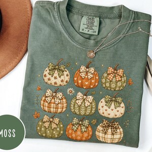 May include: A moss green short-sleeve t-shirt featuring a graphic print of nine pumpkins, each decorated with a unique patterned bow. The pumpkins display various colors and designs, including plaid, hearts, and floral motifs, accented by small leaves.