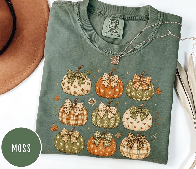 May include: A moss green short-sleeve t-shirt featuring a graphic print of nine pumpkins, each decorated with a unique patterned bow. The pumpkins display various colors and designs, including plaid, hearts, and floral motifs, accented by small leaves.