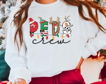May include: A white long-sleeve sweatshirt featuring a graphic print of the text PEDS in a festive holiday style, with each letter decorated as a Christmas character, and the word crew written in black cursive script below with string lights.
