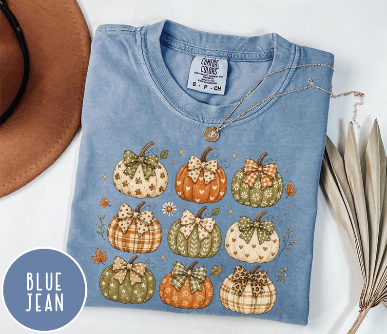May include: A blue jean colored t-shirt with a graphic print of nine pumpkins, each decorated with a patterned bow. The pumpkins feature various designs including hearts, stars, plaid, and floral patterns. The shirt has a Comfort Colors brand label.