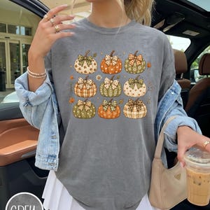 May include: A charcoal grey crewneck t-shirt featuring a graphic print of nine pumpkins arranged in a grid. Each pumpkin is decorated with a unique pattern, such as plaid, hearts, or flowers, and topped with a small bow. The shirt has a relaxed fit.