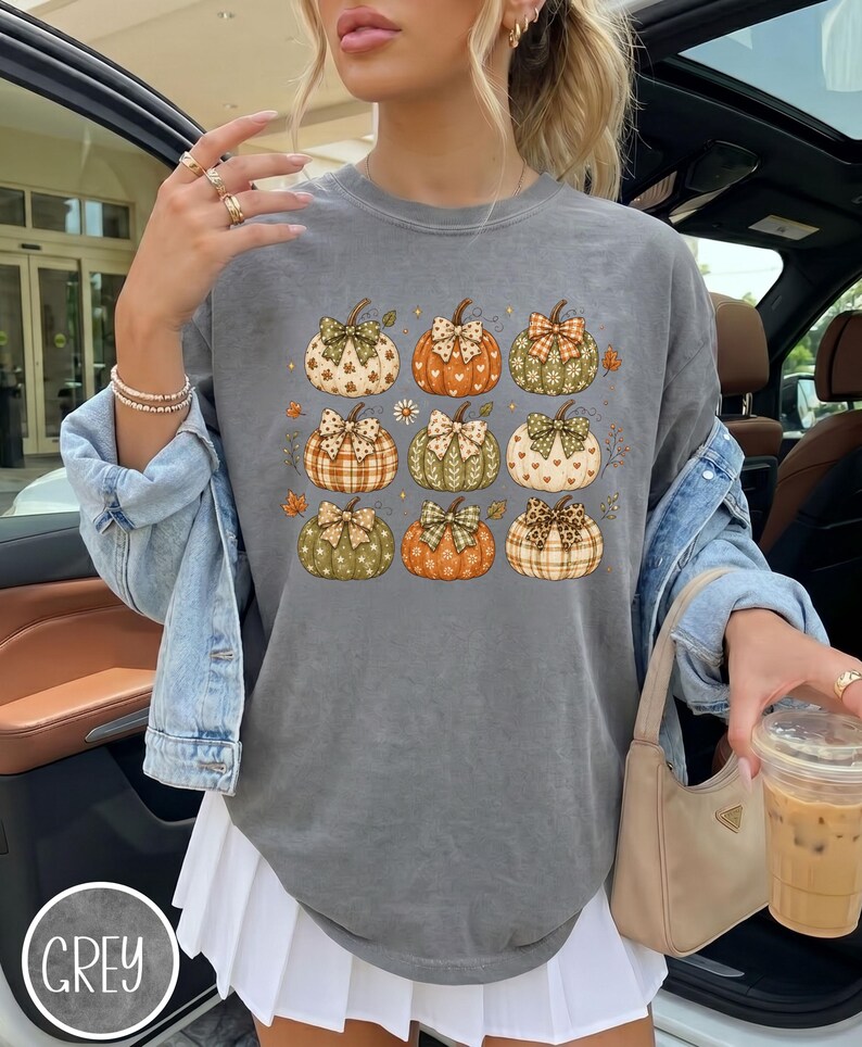 May include: A charcoal grey crewneck t-shirt featuring a graphic print of nine pumpkins arranged in a grid. Each pumpkin is decorated with a unique pattern, such as plaid, hearts, or flowers, and topped with a small bow. The shirt has a relaxed fit.