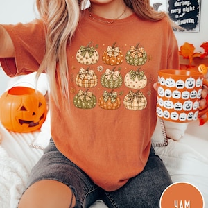 May include: A burnt orange t-shirt featuring a graphic print of nine pumpkins in various patterns, each adorned with a decorative bow. The pumpkins are arranged in a grid and display motifs including plaid, hearts, and leaves in shades of green, orange, and cream.