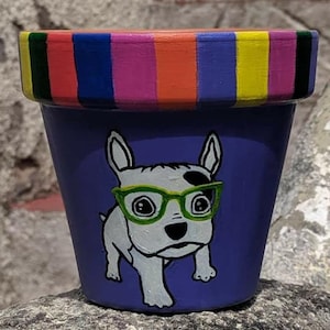 Puede incluir: Una maceta de cerámica pintada a mano con una base morada y rayas de colores alrededor de la parte superior. La maceta presenta un perro blanco con manchas negras que lleva gafas verdes.