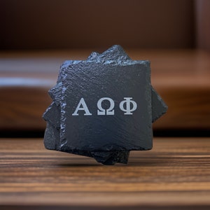 May include: A stack of black slate coasters with the Greek letters AΩΦ etched in the center. The coasters have a rough, textured surface and are displayed on a wooden surface.
