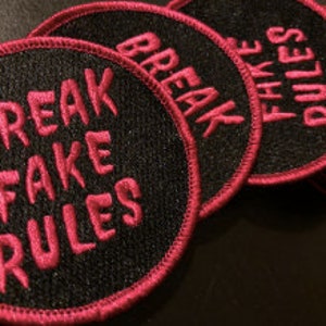 Pode incluir: Um conjunto de patches redondos bordados em preto e rosa com o texto "Break Fake Rules" em uma fonte estilizada.