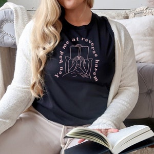 Könnte beinhalten: Eine Frau trägt ein schwarzes T-Shirt mit dem Text "You had me at reverse harem" in rosa Schrift. Sie sitzt auf einem Sofa und liest ein Buch.