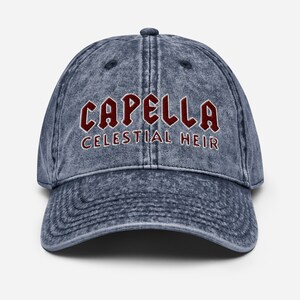 Może przedstawiać: Dżinsowa niebieska czapka bejsbolówka z wyhaftowanym słowem "CAPELLA" w kolorze bordowym, nad napisem "CELESTIAL HEIR". Czapka ma zakrzywiony daszek i guzik na górze.