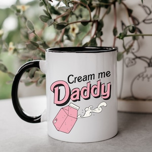 May include: White ceramic mug with a black handle and rim. The mug features the text "Cream me Daddy" in pink and black lettering, along with a pink milk carton graphic and a stream of milk.
