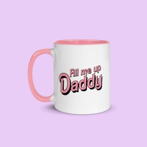 Puede incluir: Taza de cerámica blanca con borde y asa de color rosa. La taza tiene un gráfico rosa que dice "Fill me up Daddy" en una fuente retro.