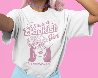 May include: White t-shirt with a pink graphic design that says "She's a Bookish Girl" and features a woman with books stacked on her head. The text below the graphic says "In a fictional world."