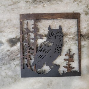 May include: A wooden wall hanging with a cutout silhouette of an owl in a forest setting. The owl is black and the forest is brown.