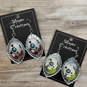 May include: Silver-toned dangle earrings with an oval, ornate frame. Each earring features a cartoon panda design with blue eyes. The earrings are displayed on black cards with the text "Slayer Creations".