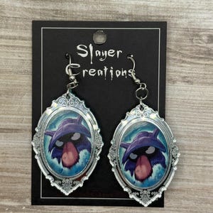 May include: A pair of silver-toned earrings featuring an oval-shaped design with a cartoon character illustration. The character has a purple hat, black eyes, and a pink tongue. The earrings are on a black card with the text "Slayer Creations".