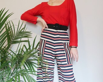 Könnte beinhalten: Eine Frau trägt ein langärmliges rotes Shirt und eine gestreifte Hose mit einem schwarzen Gürtel. Sie trägt schwarze Schuhe mit Absatz und eine goldene Halskette.