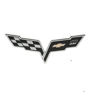 Op de afbeelding: Een zwart en zilver Chevrolet Corvette embleem met een geruit vlagontwerp en het nummer "100" in goud.