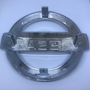 Peut inclure: Un emblème en métal argenté avec le texte "A22" en relief. L'emblème a une forme circulaire avec une section centrale rectangulaire plate.