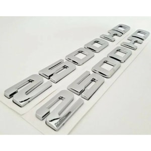 Może przedstawiać: Błyszczące, chromowane litery emblematu samochodowego z napisem "2000", ułożone w dwóch rzędach. Litery są pojedyncze i wydają się być samoprzylepne, co ułatwia aplikację. Tło to gładka, biała powierzchnia.