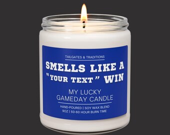 May include: A lit, white candle in a glass jar with a blue label. The label reads "Smells Like A 'Your Text' Win" and "My Lucky Gameday Candle." The candle is hand-poured with a soy wax blend and has a 50-60 hour burn time.