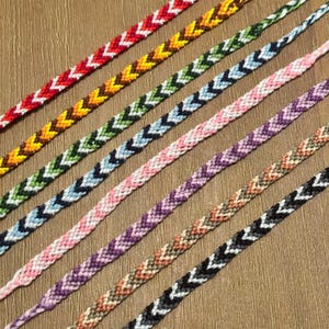Könnte beinhalten: Eine Sammlung von acht dünnen, gewebten Freundschaftsarmbändern, die diagonal angeordnet sind. Jedes Armband weist ein ausgeprägtes Zickzackmuster aus zweifarbigem Stickgarn auf, darunter Rot und Weiß, Gelb und Braun, Grün und Weiß, Blau und Weiß, Rosa und Weiß, Violett und Weiß, Orange und Beige sowie Schwarz und Weiß.