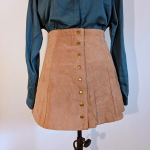 May include: A tan suede mini skirt with a button-down front. The skirt has a high waist and a slight A-line shape. The buttons are gold-coloured. The skirt is displayed on a black mannequin stand, paired with a teal button-down shirt.