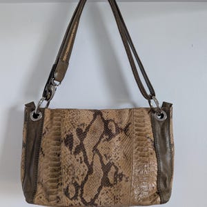 May include: A brown and tan snakeskin-patterned handbag with dark brown leather accents and straps. The bag has silver-coloured metal rings connecting the straps to the body. The bag is rectangular in shape.
