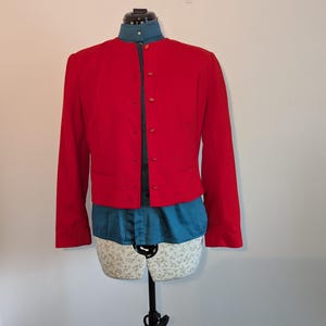 May include: A vibrant red jacket with a mandarin collar and long sleeves. The jacket is fastened with buttons and worn over a teal shirt. The jacket is displayed on a mannequin.