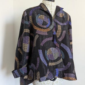 May include: A black jacket with a distinctive geometric pattern in shades of purple, blue, and tan. The jacket has a collared neckline, long sleeves, and a button closure. The fabric has a textured appearance, and the design is striking.