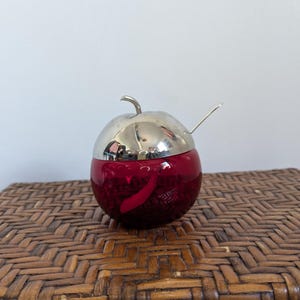May include: A red apple-shaped container with a silver lid and spoon. The container is made of glass and has a shiny, reflective surface. The lid is also silver and has a stem-like handle. The spoon is small and silver.