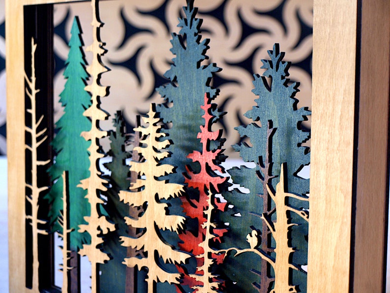 May include: A wooden wall art piece featuring a layered forest scene with trees in shades of green, brown, and red. The trees are cut out and layered to create a 3D effect.