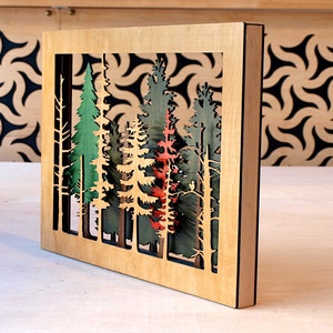 May include: A wooden wall art piece with a cutout design of a forest scene. The trees are painted in shades of green, red, and brown. The piece is framed in a natural wood frame.