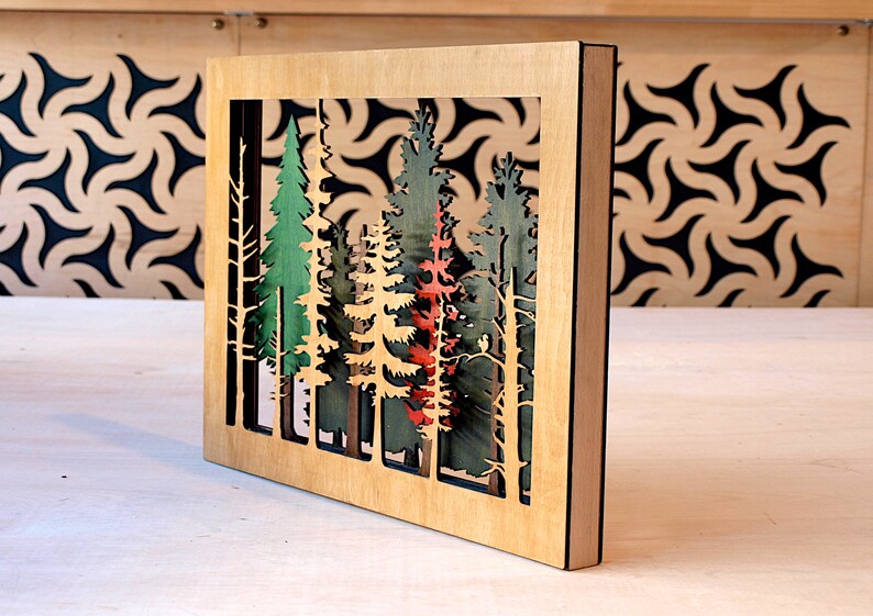 May include: A wooden wall art piece with a cutout design of a forest scene. The trees are painted in shades of green, red, and brown. The piece is framed in a natural wood frame.