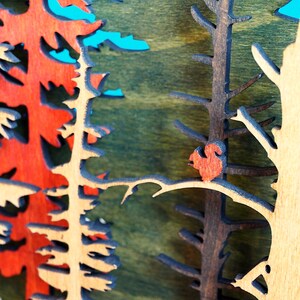 May include: A wooden wall art piece depicting a forest scene with a red bird perched on a branch. The trees are cut out of wood and painted in shades of brown, green, and red.