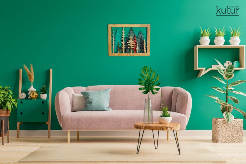 May include: A pink sofa with a blue and white throw pillow sits in a living room with a green wall. A wooden wall art piece with a forest scene hangs above the sofa. A small wooden table with a black metal frame sits in front of the sofa. A potted plant with large green leaves sits on the table. A wooden shelf with three potted plants sits on the wall to the right of the sofa.