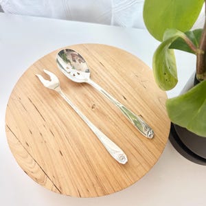 May include: A silver-toned serving spoon and a matching two-pronged serving fork. The spoon features a perforated bowl with a decorative cutout pattern, and both utensils have floral embossed detailing on the handles with a polished finish.