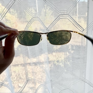 Peut inclure: Lunettes de soleil &agrave; monture en m&eacute;tal dor&eacute; dot&eacute;es de verres rectangulaires vert fonc&eacute;. Le verre droit pr&eacute;sente un petit logo blanc. La fine monture m&eacute;tallique comprend des plaquettes r&eacute;glables et des branches fines aux extr&eacute;mit&eacute;s sombres.
