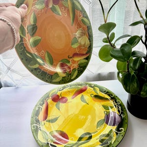 May include: Two round ceramic plates featuring a hand-painted border of assorted vegetables and fruits including garlic, peppers, and eggplant. One plate has an orange center, while the other has a yellow center, both with green rims.
