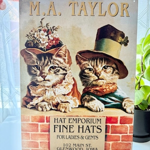 May include: A rectangular metal sign featuring two tabby cats wearing hats and Victorian-style clothing. The text reads M.A. TAYLOR, HAT EMPORIUM FINE HATS FOR LADIES & GENTS, 102 MAIN ST. GLENWOOD, IOWA TEL 28. The cats are positioned behind a red brick wall.