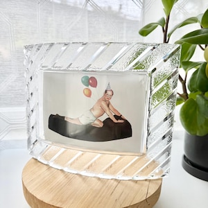 May include: A rectangular glass picture frame with a textured, diagonal striped border. Inside the frame is a print of a person wearing a party hat and white shorts, kneeling on a black surface while holding three colorful balloons in the air.