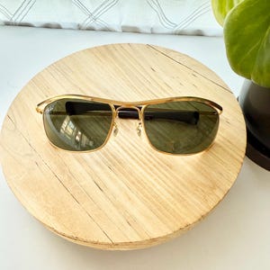 Peut inclure: Lunettes de soleil en m&eacute;tal dor&eacute; dot&eacute;es d'une monture rectangulaire avec un double pont et des verres teint&eacute;s vert fonc&eacute;. Les branches sont termin&eacute;es par des embouts en plastique noir, offrant une structure classique avec des d&eacute;tails m&eacute;talliques.