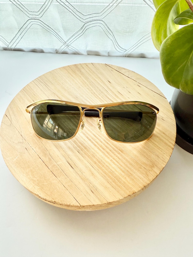 Peut inclure: Lunettes de soleil en m&eacute;tal dor&eacute; dot&eacute;es d'une monture rectangulaire avec un double pont et des verres teint&eacute;s vert fonc&eacute;. Les branches sont termin&eacute;es par des embouts en plastique noir, offrant une structure classique avec des d&eacute;tails m&eacute;talliques.