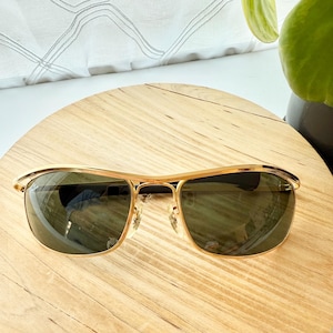 Peut inclure: Lunettes de soleil dor&eacute;es dot&eacute;es d'une barre frontale incurv&eacute;e et &eacute;l&eacute;gante ainsi que de verres rectangulaires teint&eacute;s en vert fonc&eacute;. La monture pr&eacute;sente une finition m&eacute;tallique polie, des plaquettes r&eacute;glables et des branches fines et lisses.